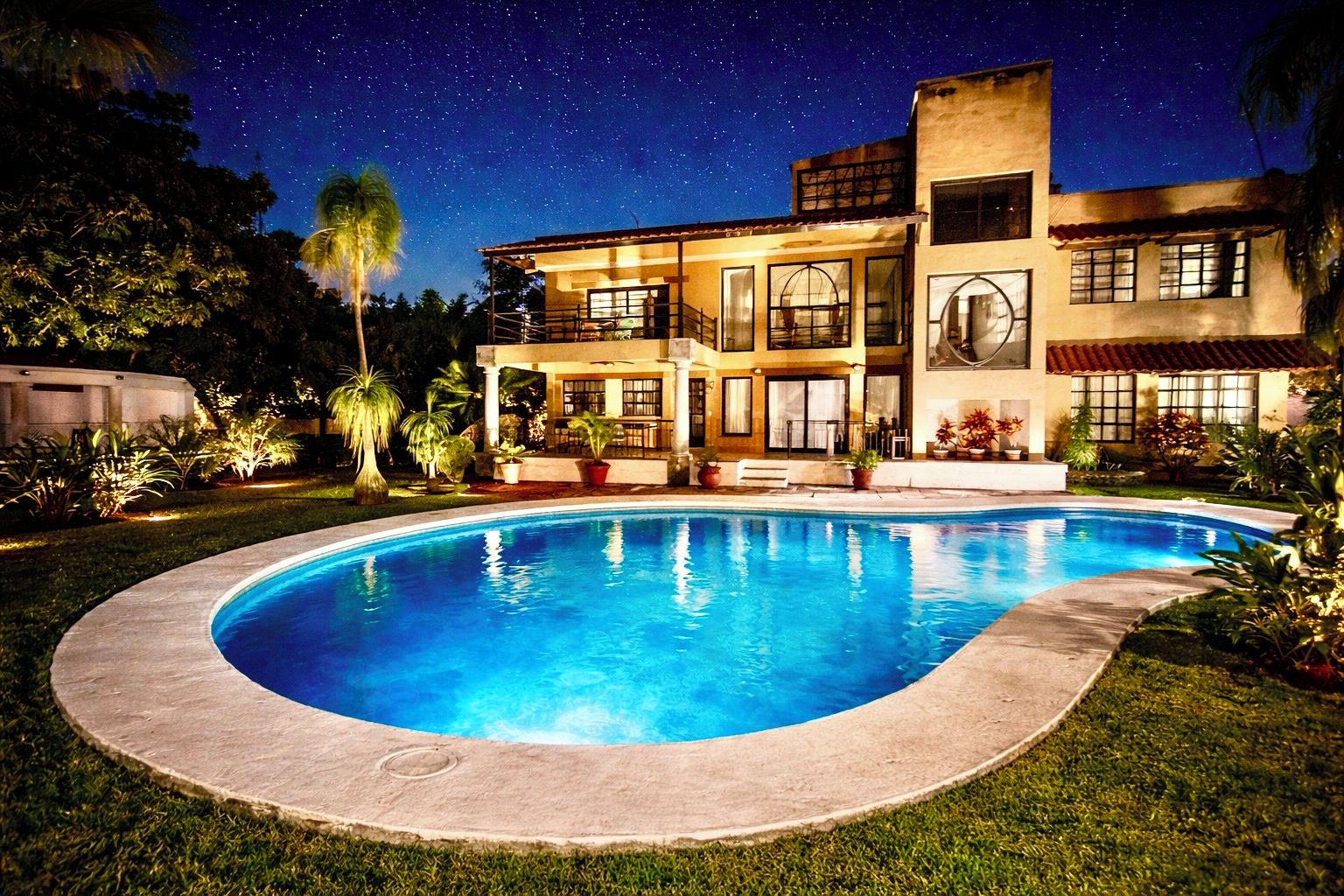 Una casa de dos pisos con piscina, rodeada de follaje bajo un cielo estrellado.