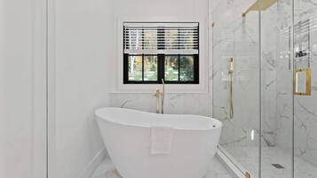 A modern bathroom with a freestanding bathtub, a shower enclosure, and a window with blinds.