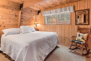 A wooden cabin bedroom with a bed, a rocking chair, a nightstand, and a window with a view of trees. - Zen Den (Big Bear Lake)
