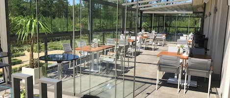 Un patio exterior con paredes de vidrio, mesas, sillas y un pérgola.