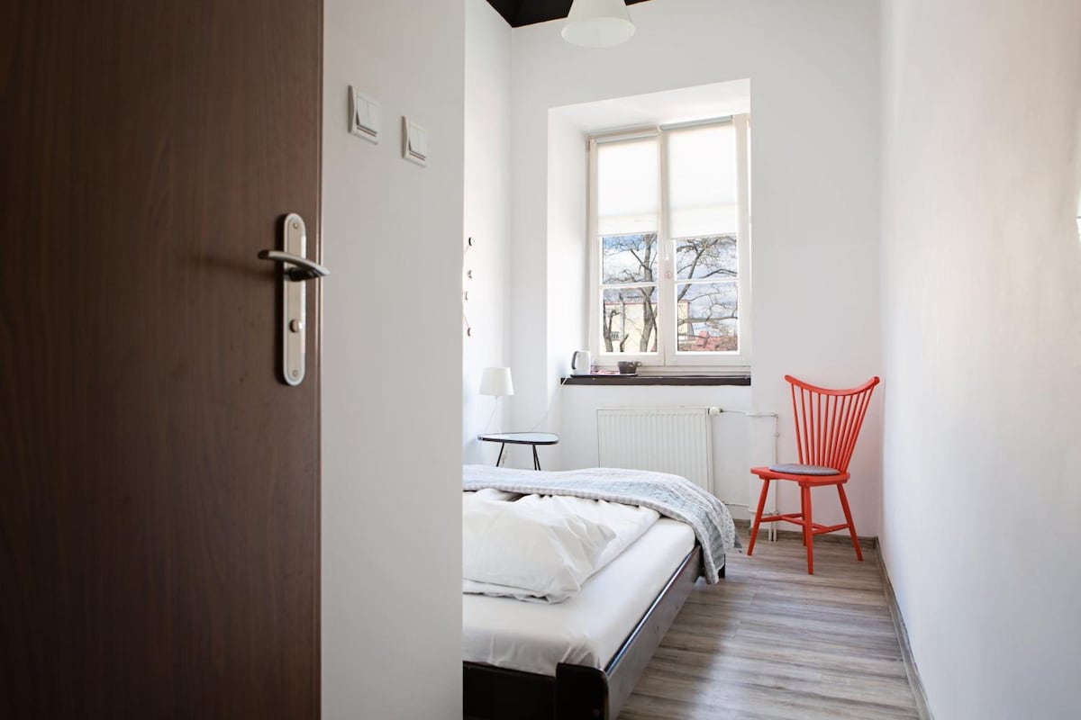 A bedroom with a bed, a window, a radiator, a chair, and a door.