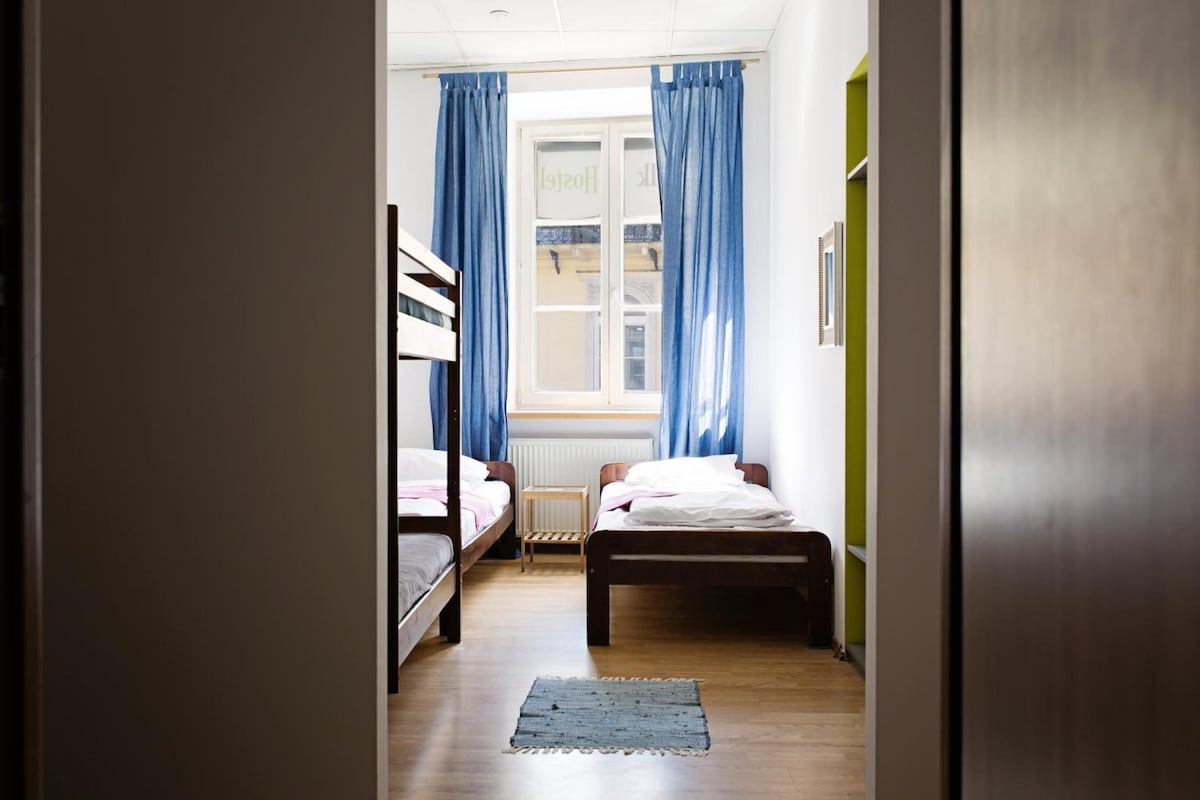 A dormitory room with bunk beds, a window with curtains, a radiator, and a small table.