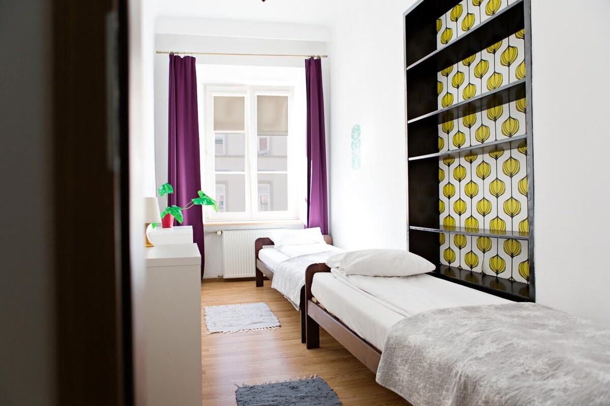 A bedroom with two beds, a window with purple curtains, a radiator, and a shelf with patterned wallpaper.