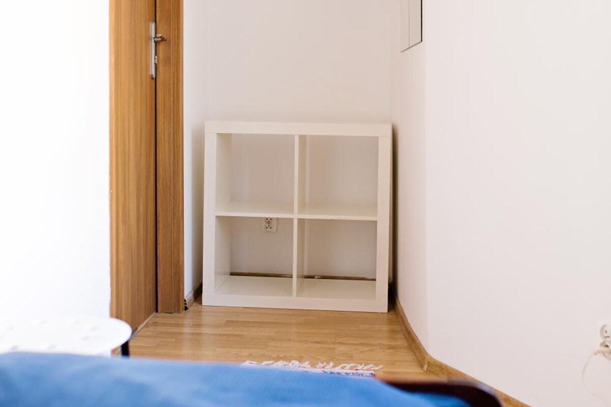 A narrow hallway with a wooden door on the left, a white shelf unit in the center, and a blue bed at the end.
