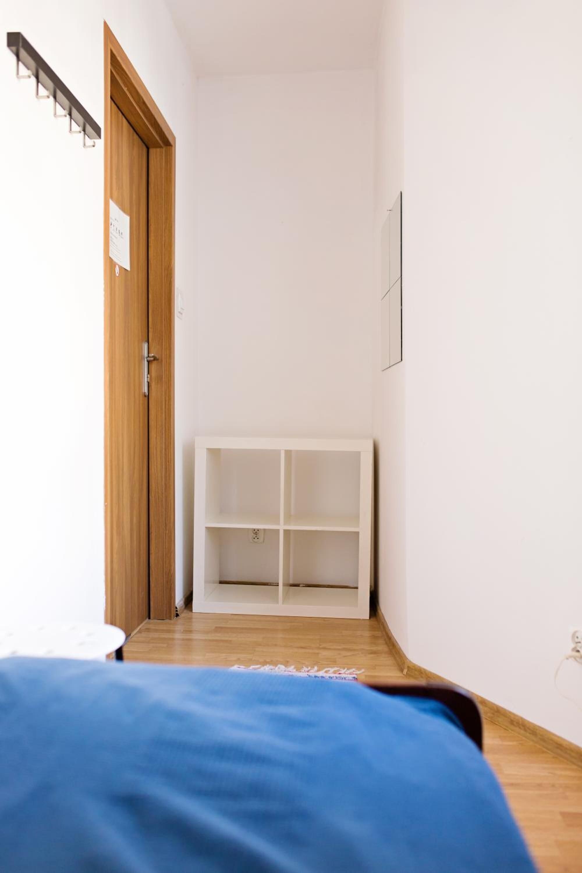 Un pasillo estrecho con una puerta de madera a la izquierda, un estante blanco en el centro y una cama azul al final.