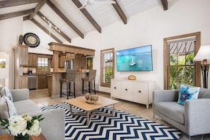 Amplia sala de estar con cocina, comedor con sillas de mimbre, televisor de pantalla plana y sofá con cojines decorativos.