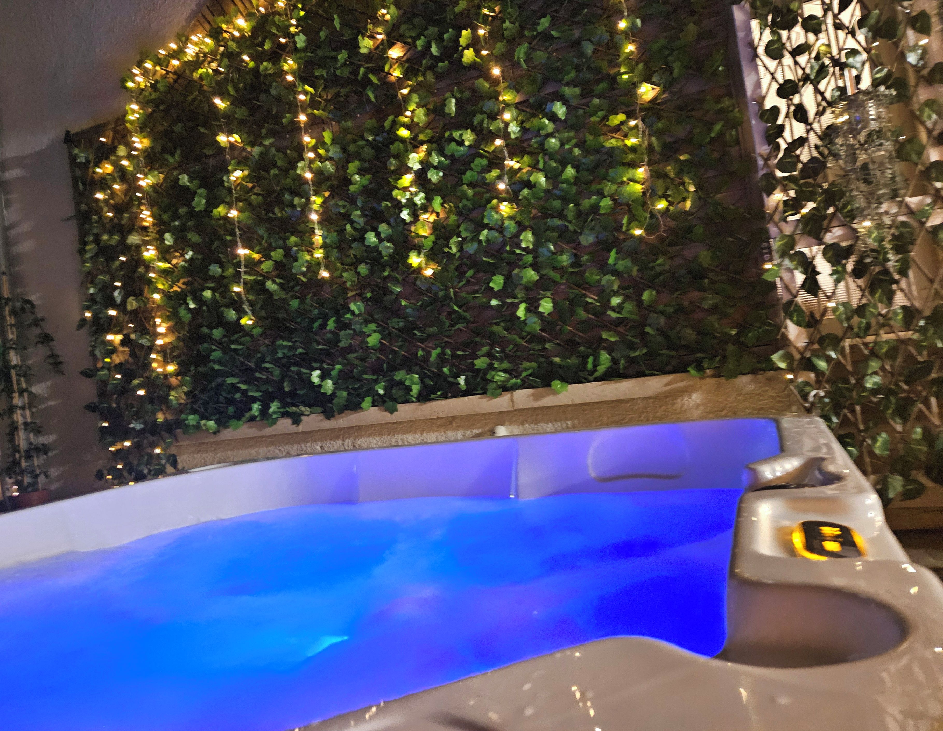 Un jacuzzi con iluminación azul en su interior, rodeado de follaje y luces colgantes.