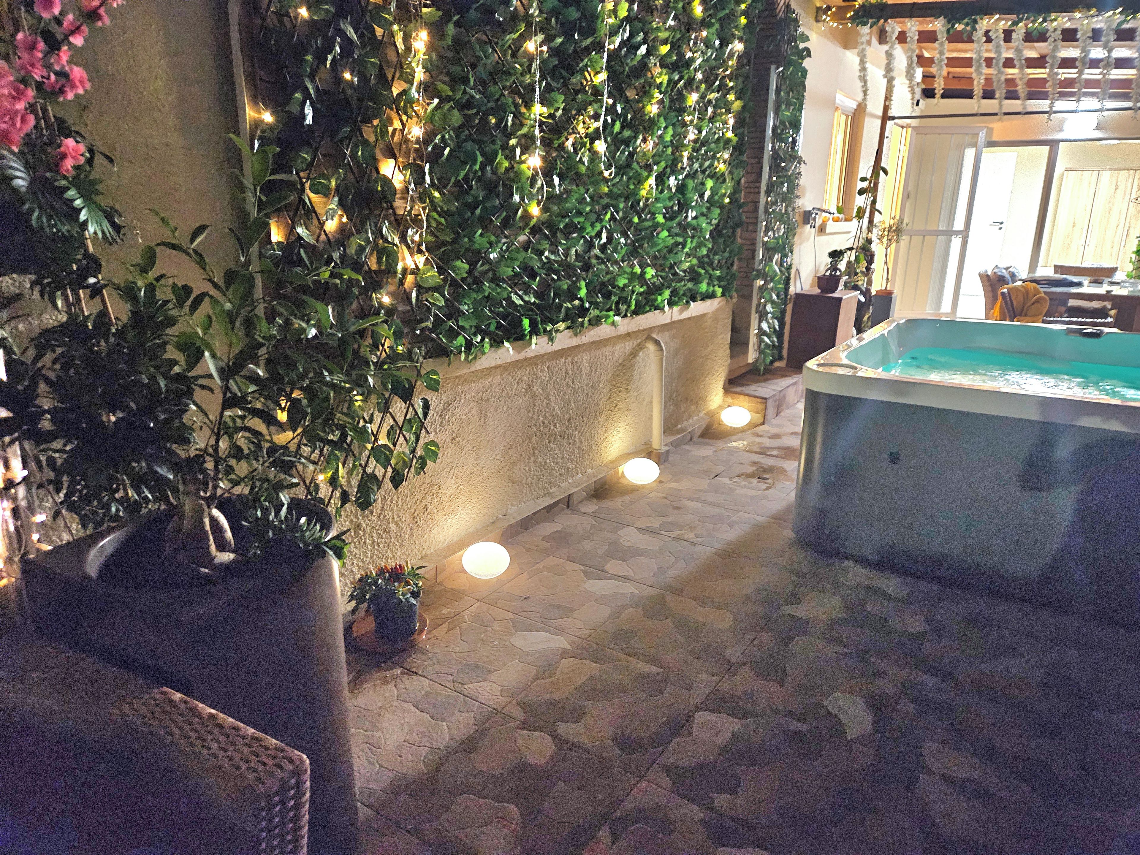 An outdoor area with a stone floor, a hot tub, and a wall covered with greenery and string lights.