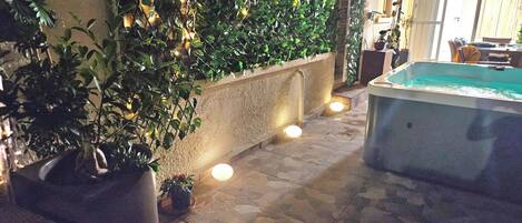 An outdoor area with a stone floor, a hot tub, and a wall covered with greenery and string lights.