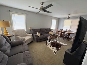 A living room with a grey sofa, a brown leather ottoman, a dining table with chairs, a ceiling fan, and a television. - Beautiful Home for a Relaxing Stay (Phenix City)