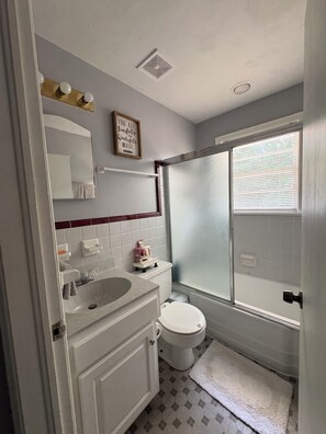 A bathroom with a white sink, mirror, and toilet. There is a shower with a glass door and a window above it. - Beautiful Home for a Relaxing Stay (Phenix City)
