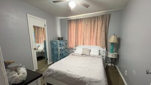 Un dormitorio con cama, cómoda, mesita de noche con lámpara, ventilador de techo y puerta que da a otra habitación.