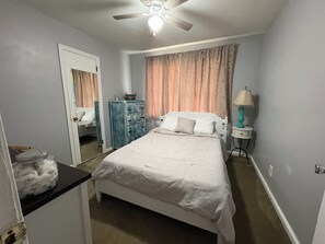 A bedroom with a bed, a dresser, a nightstand with a lamp, a ceiling fan, and a door leading to another room. - Beautiful Home for a Relaxing Stay (Phenix City)
