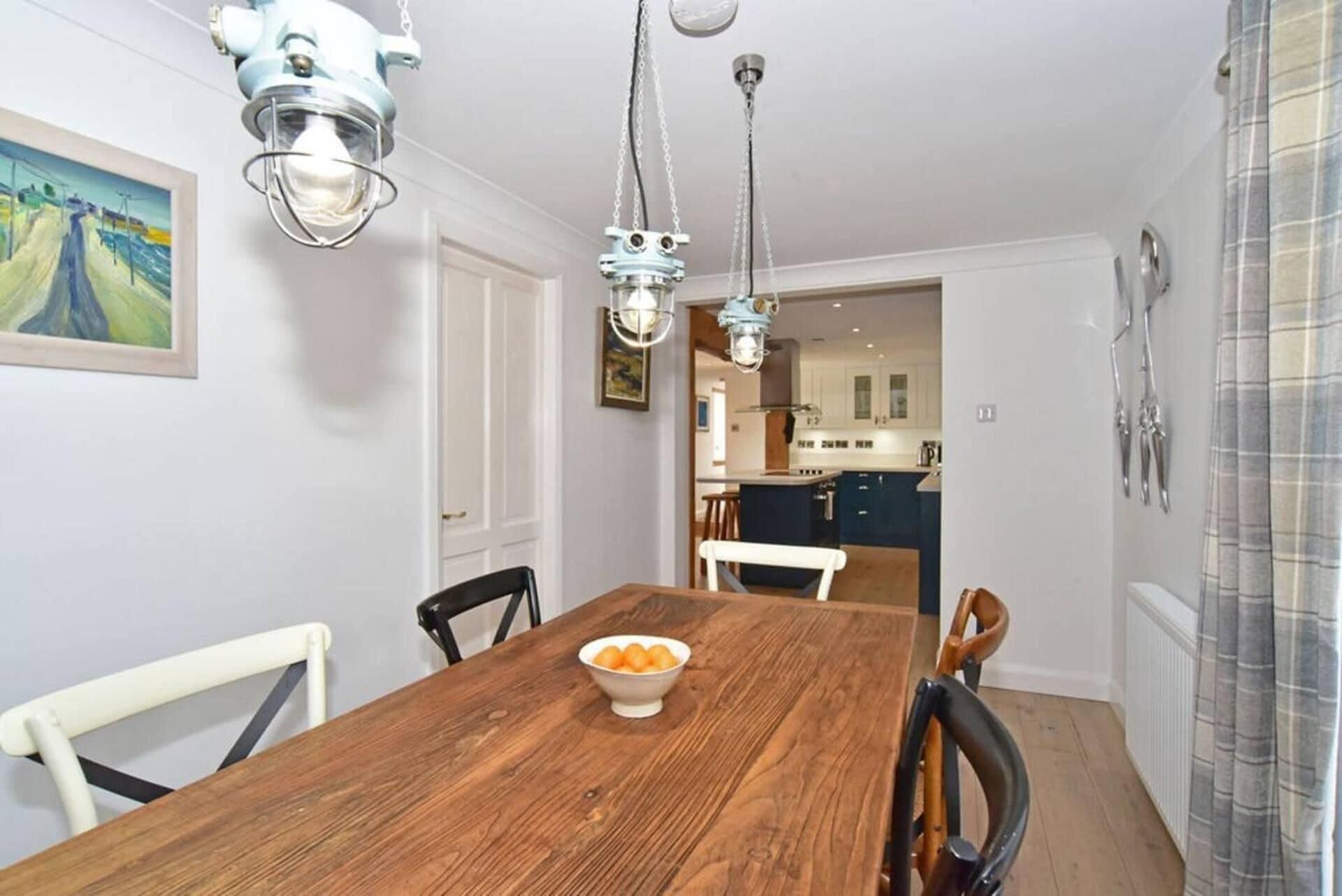 A dining area with a wooden table, chairs, and a bowl of fruit. There is a kitchen in the background with modern appliances and a view of the outside.