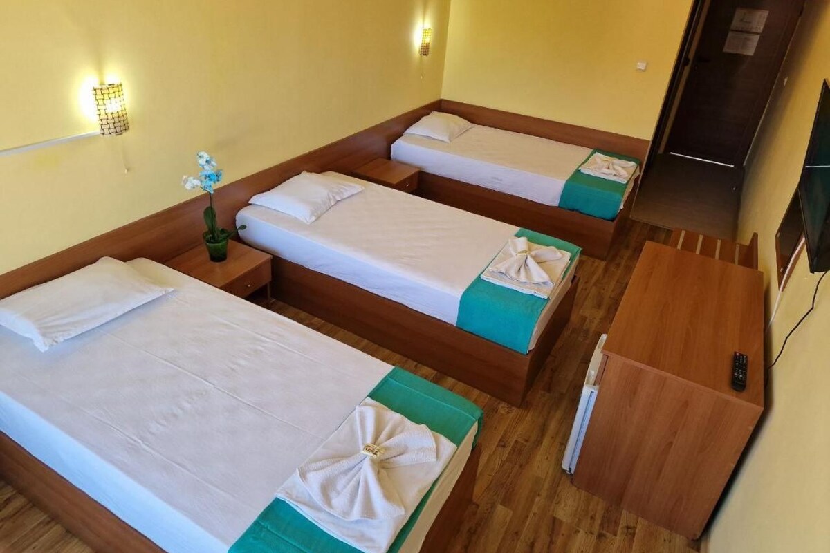 A hotel room with three single beds, a wooden desk, a small table, and a wall-mounted TV.