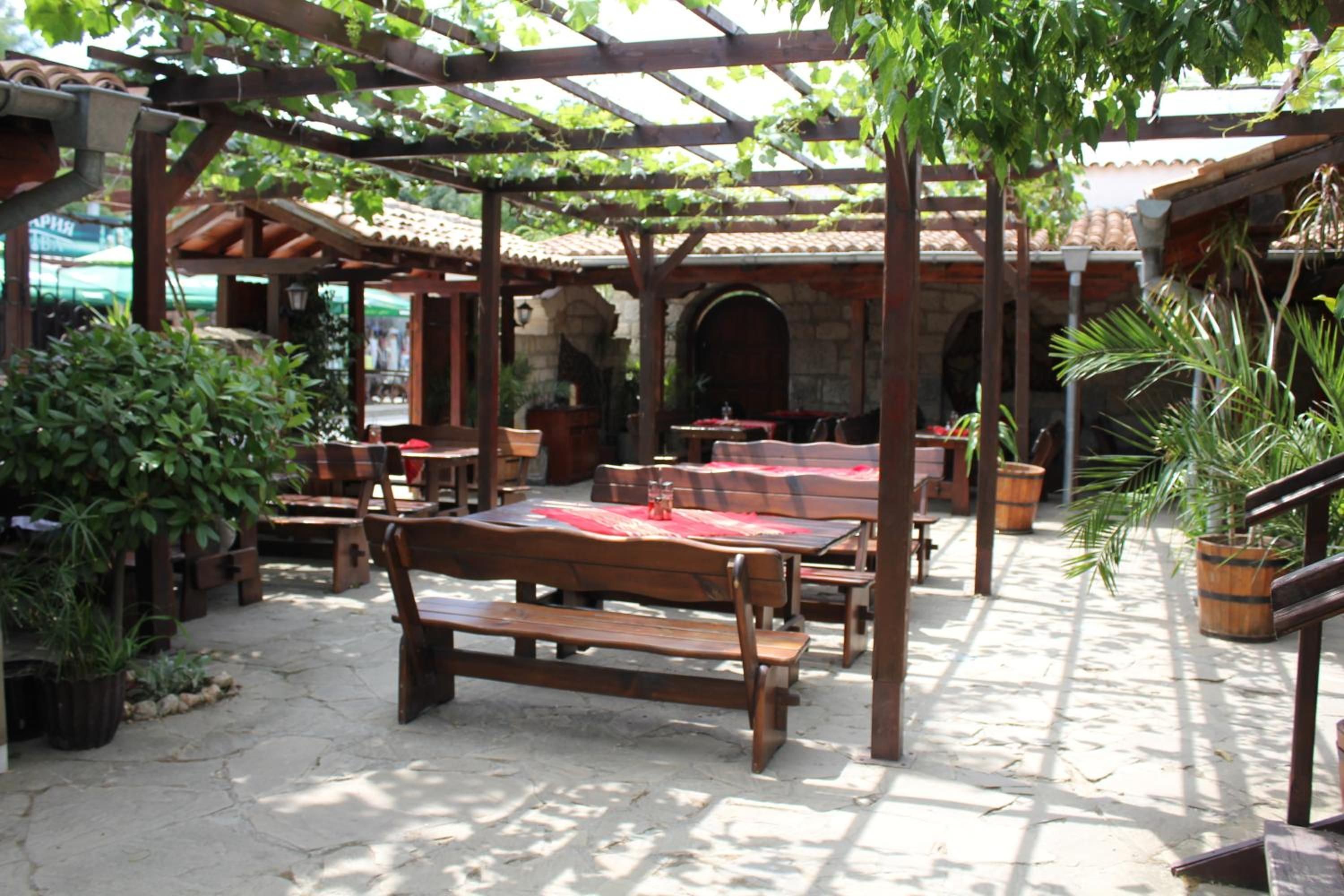 Un patio al aire libre con mesas y bancos de madera, rodeado de plantas en macetas y una estructura de pérgola.