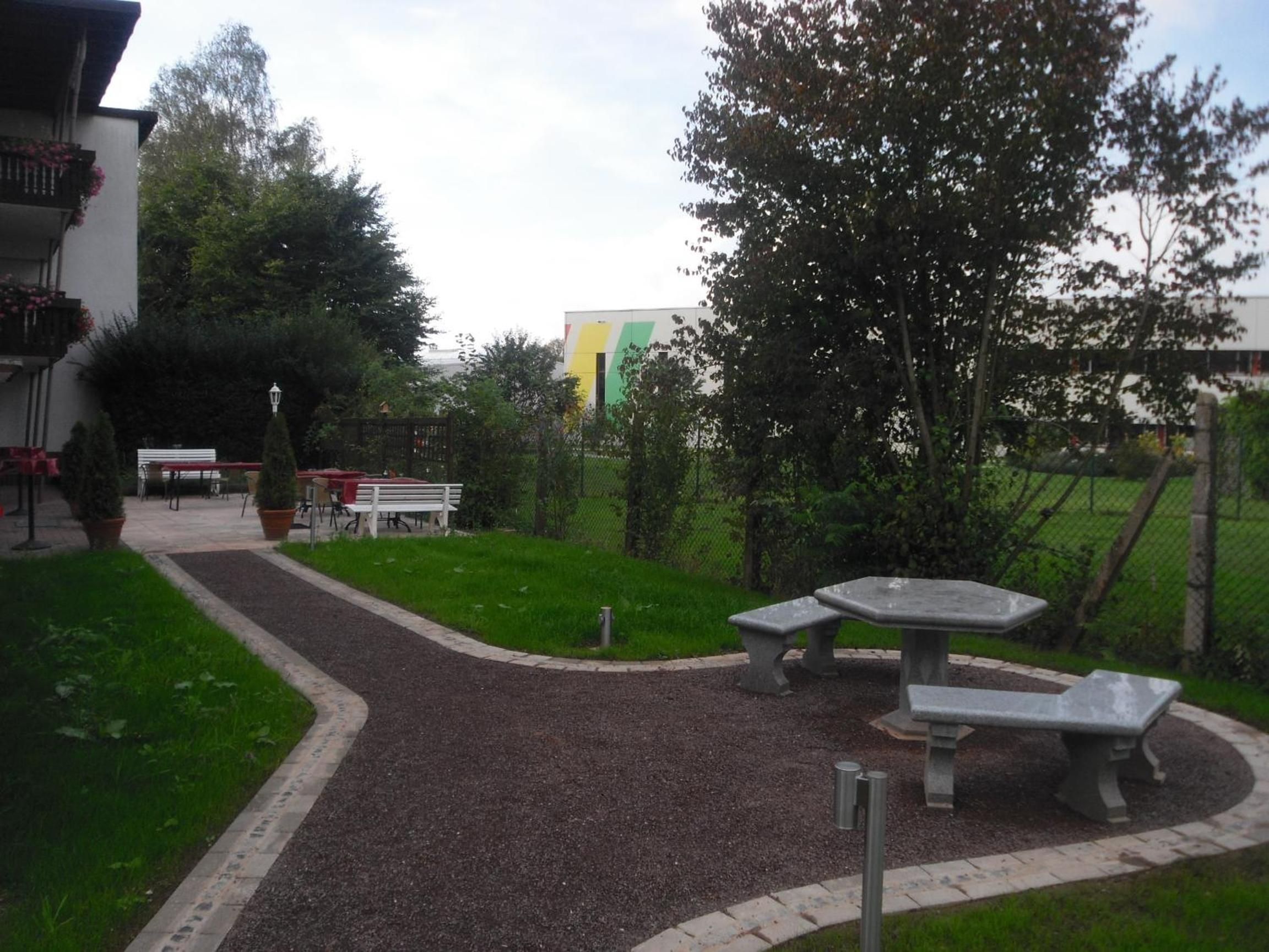 A gravel pathway leading to a seating area with benches and a table, surrounded by greenery and a fence.