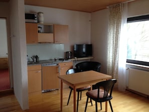 Een kleine keuken met een magnetron, waterkoker en een televisie op een plank. Er staat een eettafel met twee stoelen en een raam met gordijnen.