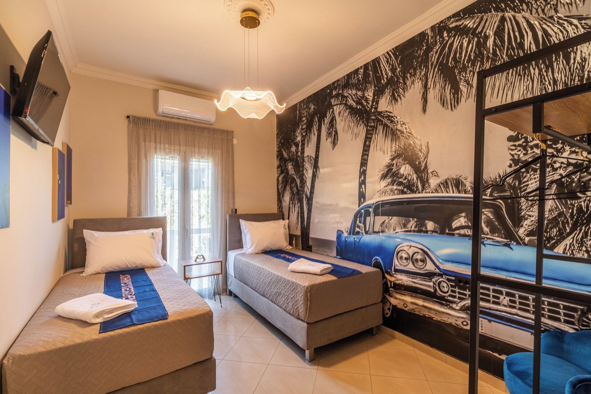 Een hotelkamer met twee bedden, een groot raam, een airconditioning, een tv aan de muur, en een muurschildering van een auto en palmbomen.