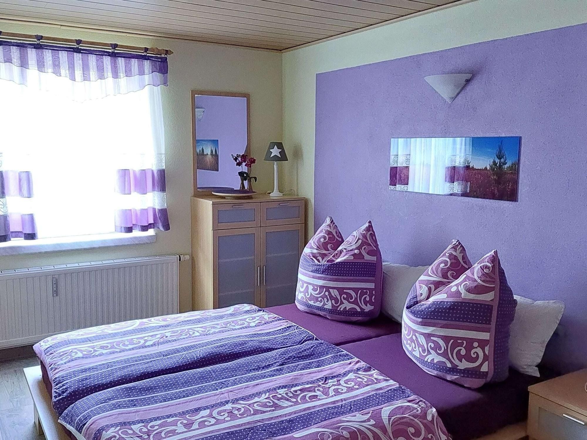 Een kamer met een bed, twee kussens, een nachtkastje, een spiegel en een raam met gordijnen.