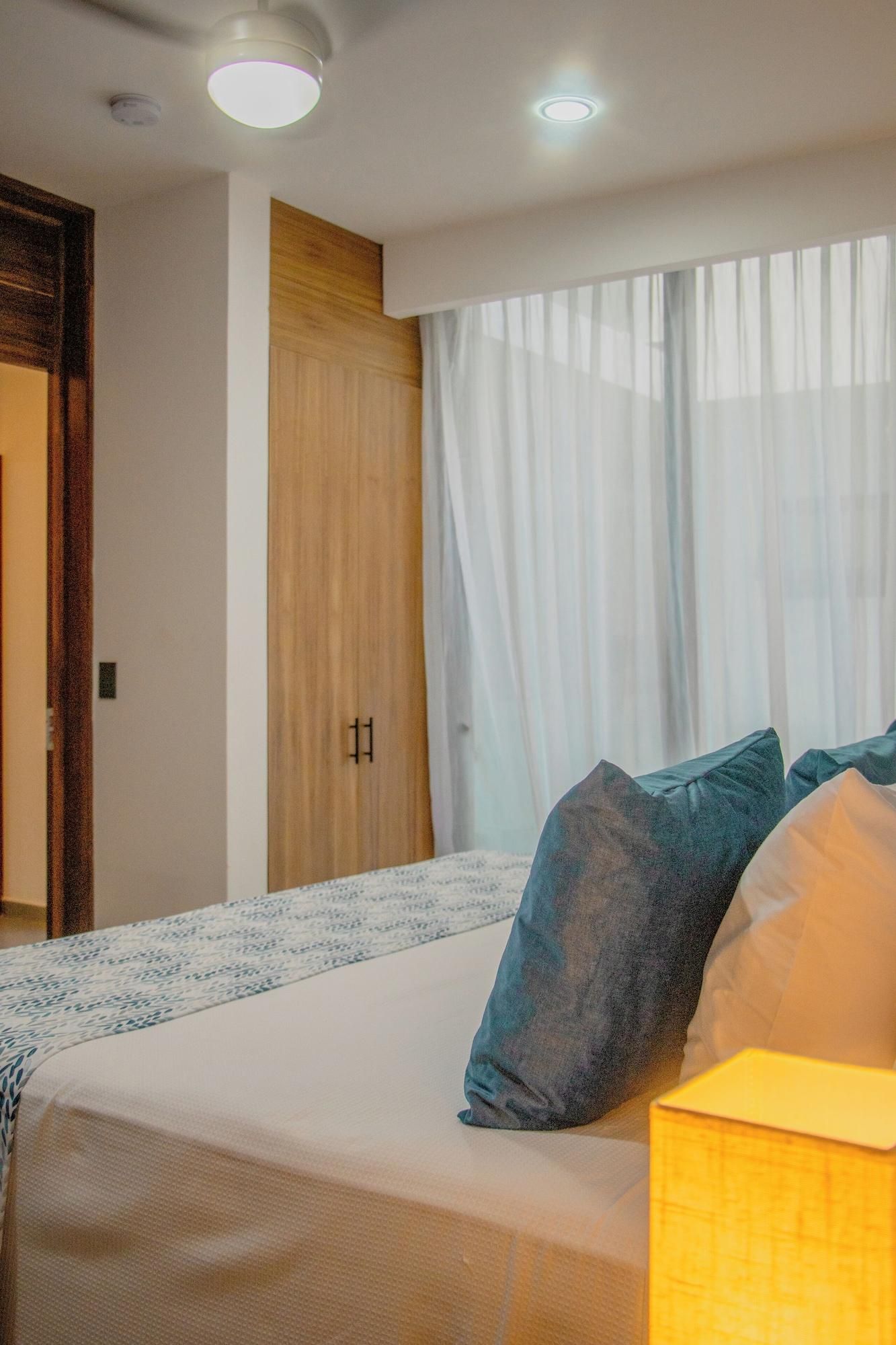 Habitación de hotel con cama, mesita de noche, armario de madera y ventana con cortinas translúcidas.