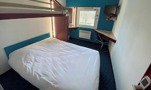 A bunk bed with a white mattress, a desk with a chair, a window with a view of trees, and a television on the wall.