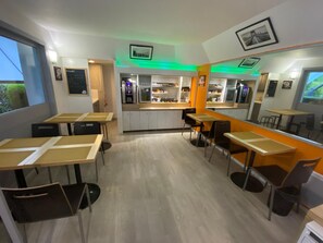 A modern cafe with multiple tables and chairs, a counter with a refrigerator and coffee machines, and a view of the outdoors through large windows.