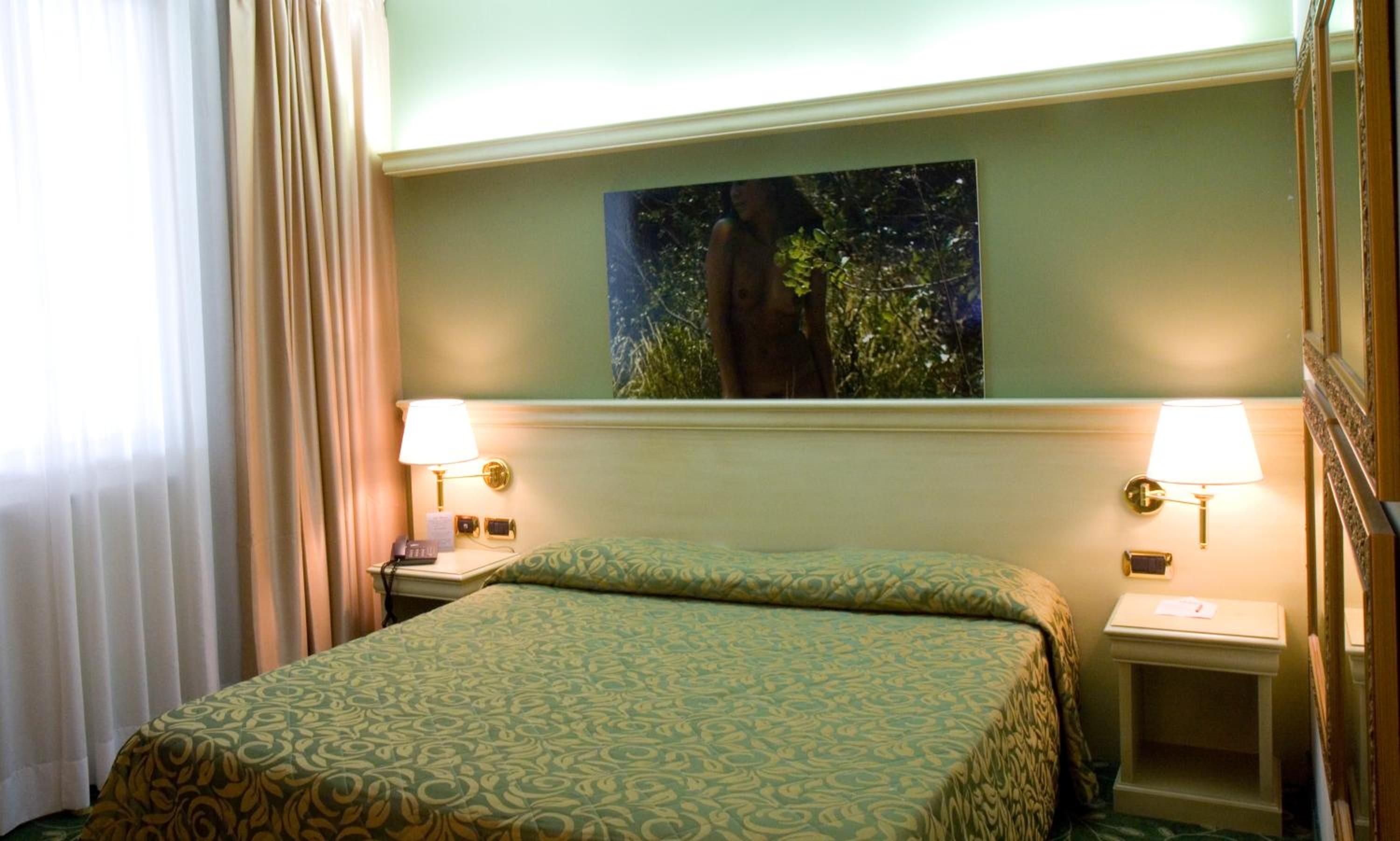 Ein Hotelzimmer mit einem Bett, zwei Nachttischlampen, einem Nachttisch, einem Bild an der Wand und einem Fenster mit Vorhängen.