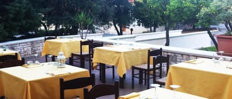An outdoor dining area with tables covered in yellow tablecloths, chairs, and a stone wall in the background.