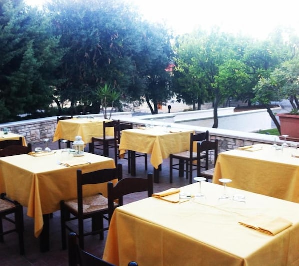 Un área de comedor al aire libre con mesas cubiertas con manteles amarillos, sillas y una pared de piedra al fondo.