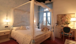 A four-poster canopy bed with a bedside table and lamp, a desk with a chair, and a large floral painting on the wall. - Villa Baroni (Bodio Lomnago)