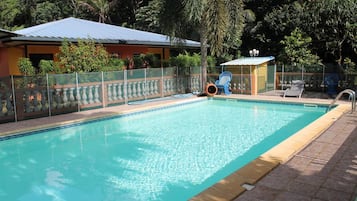 Una piscina con solárium empedrado, rodeada por una valla y abundante vegetación.