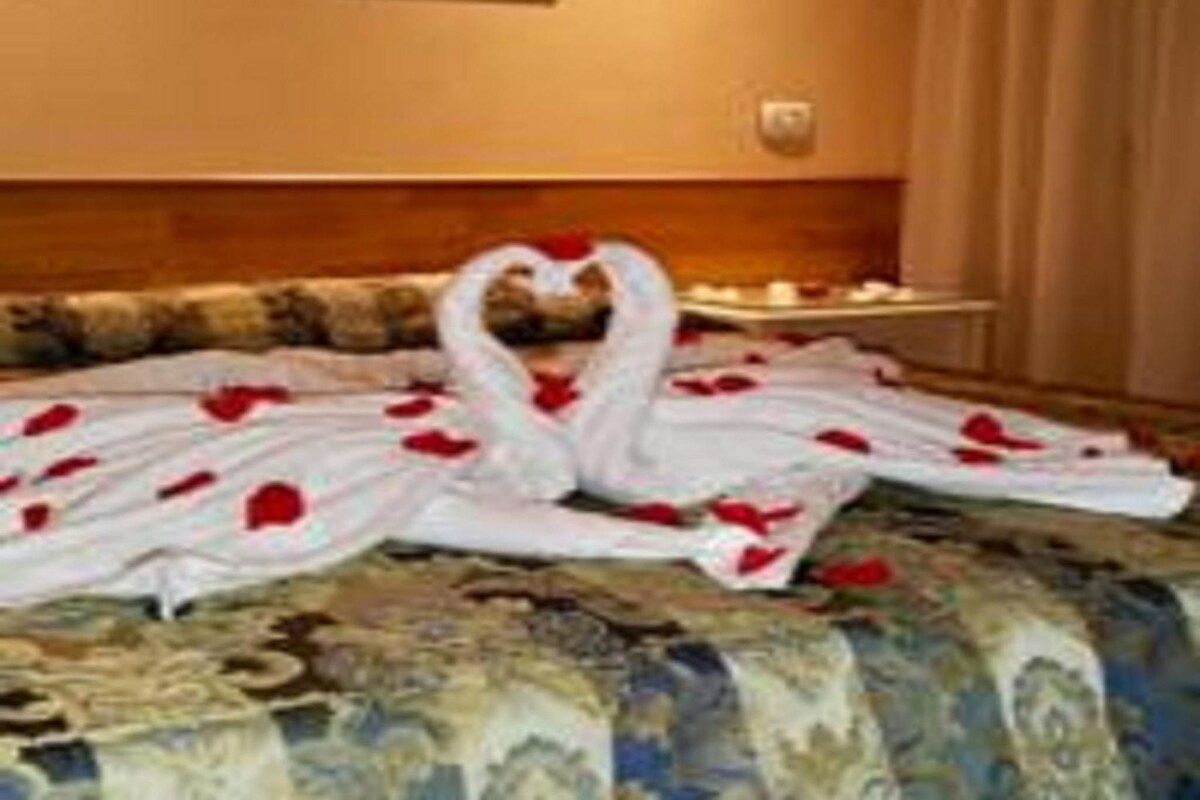 A bed with a heart-shaped arrangement of white bathrobes and red rose petals.