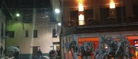 Zona exterior nevada con plantas en macetas y un edificio con iluminación cálida en su interior.