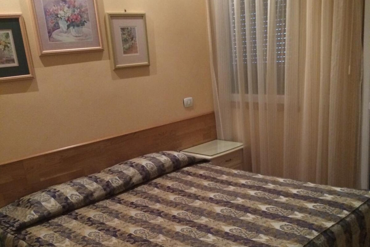 A bedroom with a bed, a nightstand, a wall-mounted light, and a window with sheer curtains.