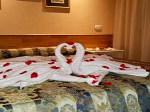 A bed with a heart-shaped arrangement of white bathrobes and red rose petals.