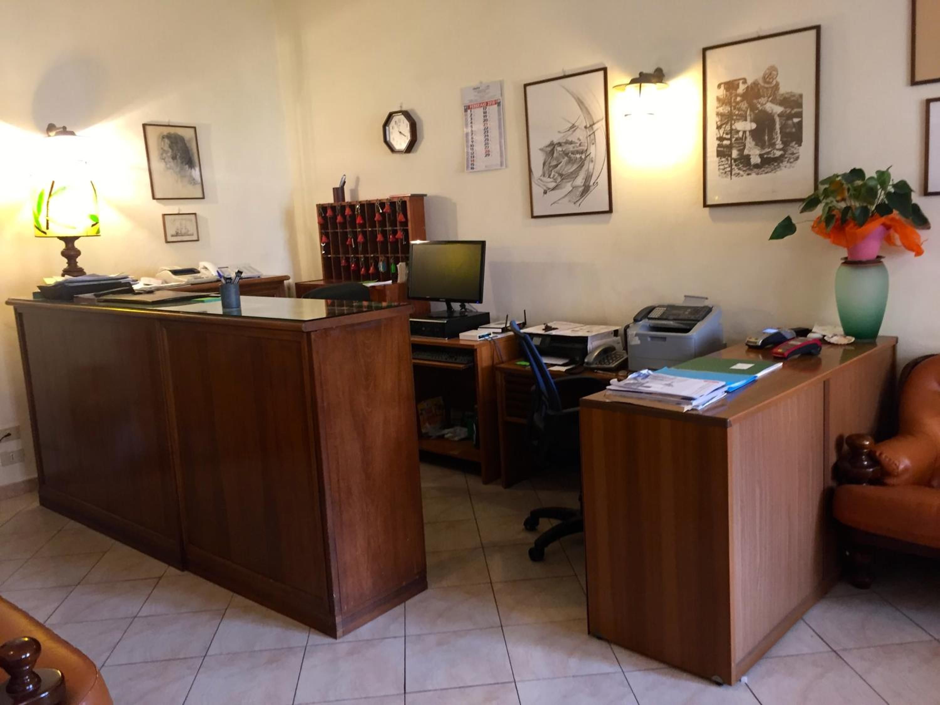 A reception desk with a computer, printer, and various office supplies. There are framed artworks on the wall and a clock.