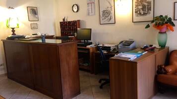 A reception desk with a computer, printer, and various office supplies. There are framed artworks on the wall and a clock.