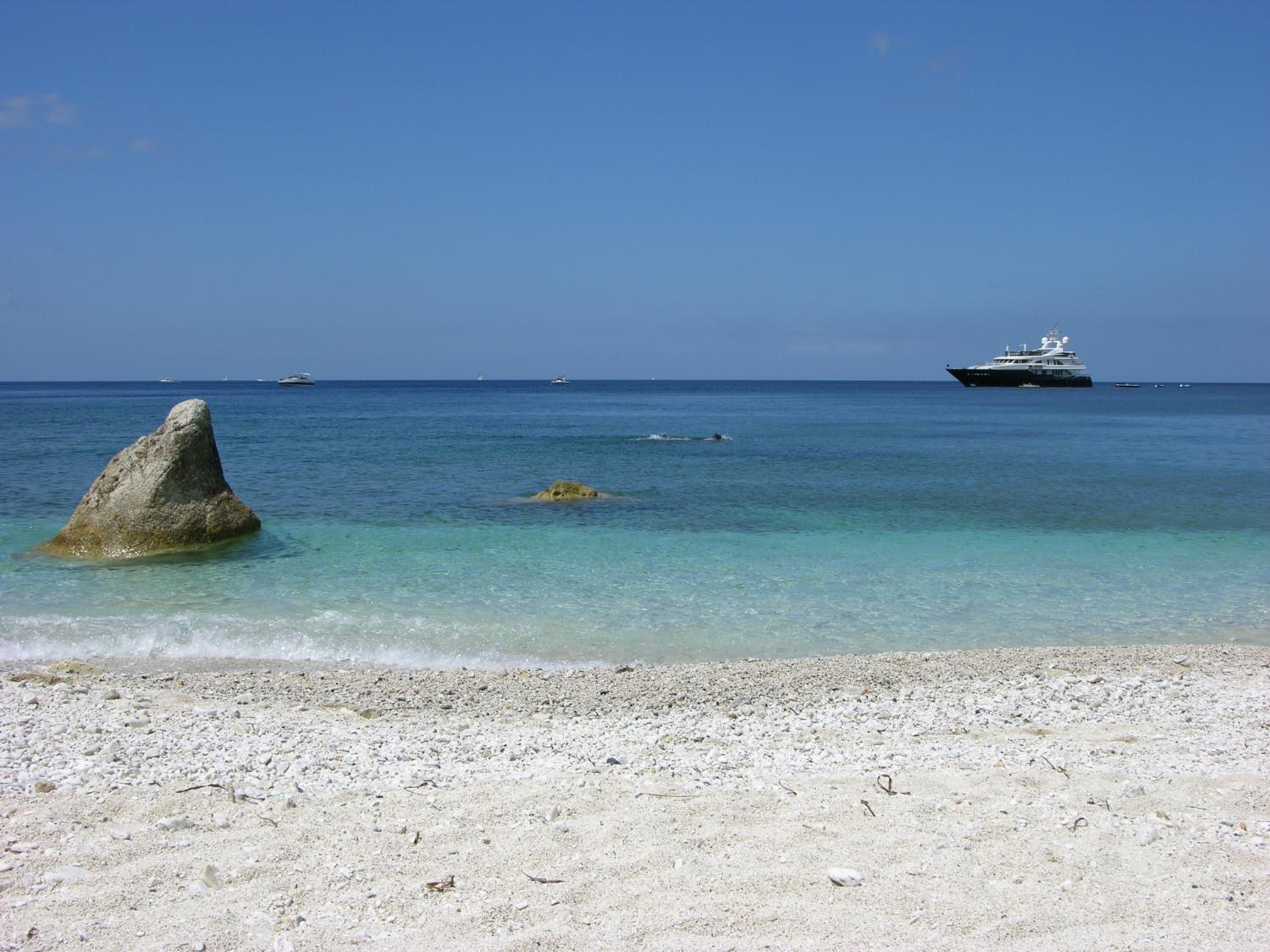 A beach with clear turquoise water, a large rock in the foreground, and a yacht in the distance.