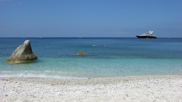 A beach with clear turquoise water, a large rock in the foreground, and a yacht in the distance.