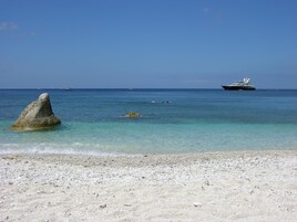 A beach with clear turquoise water, a large rock in the foreground, and a yacht in the distance.