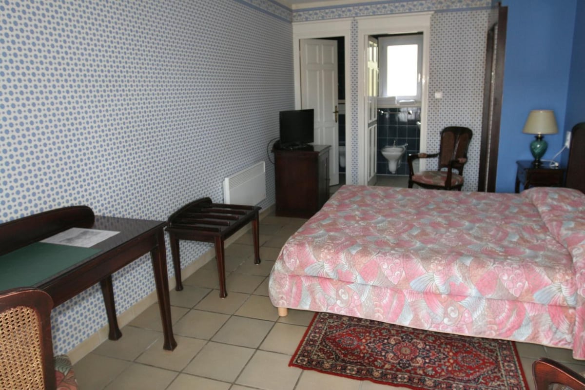 Habitación de hotel con cama, escritorio, silla, televisión y baño visible a través de una puerta abierta.