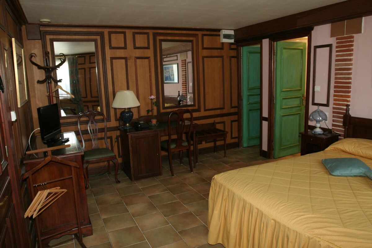 A hotel room with a bed, desk, chairs, a mirror, and a green door.