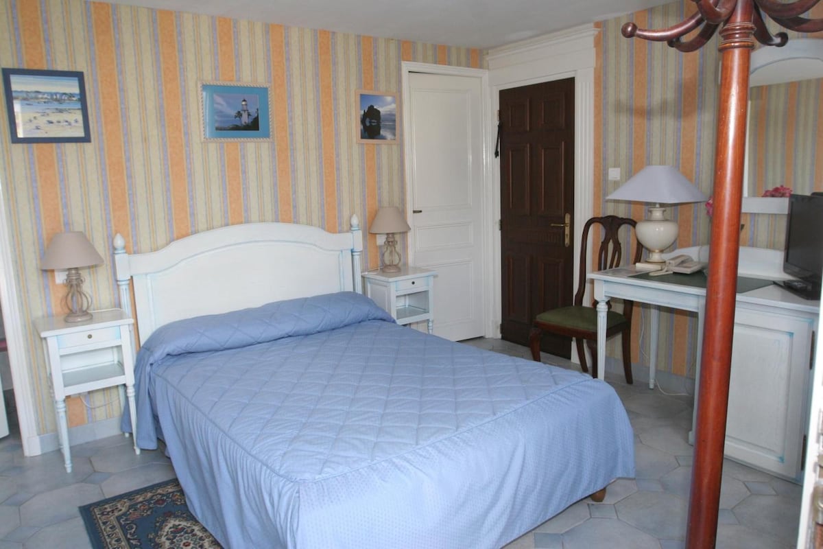 Un dormitorio con cama, mesitas de noche, una silla, un escritorio con lámpara y un televisor.