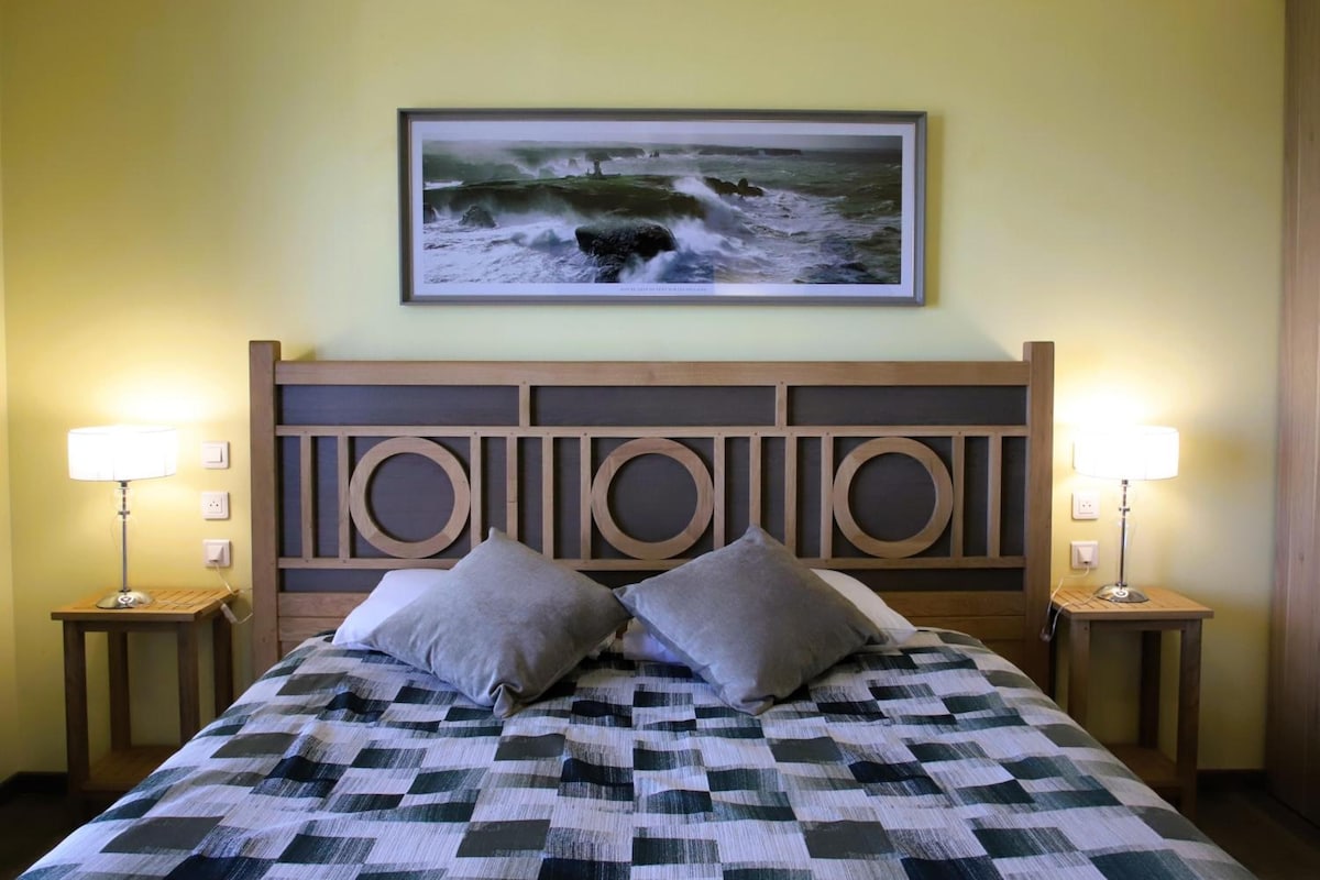 Una cama con colcha a cuadros, dos almohadas, un cabecero de madera con recortes circulares, dos lámparas de noche y un cuadro con una vista marina en la pared.