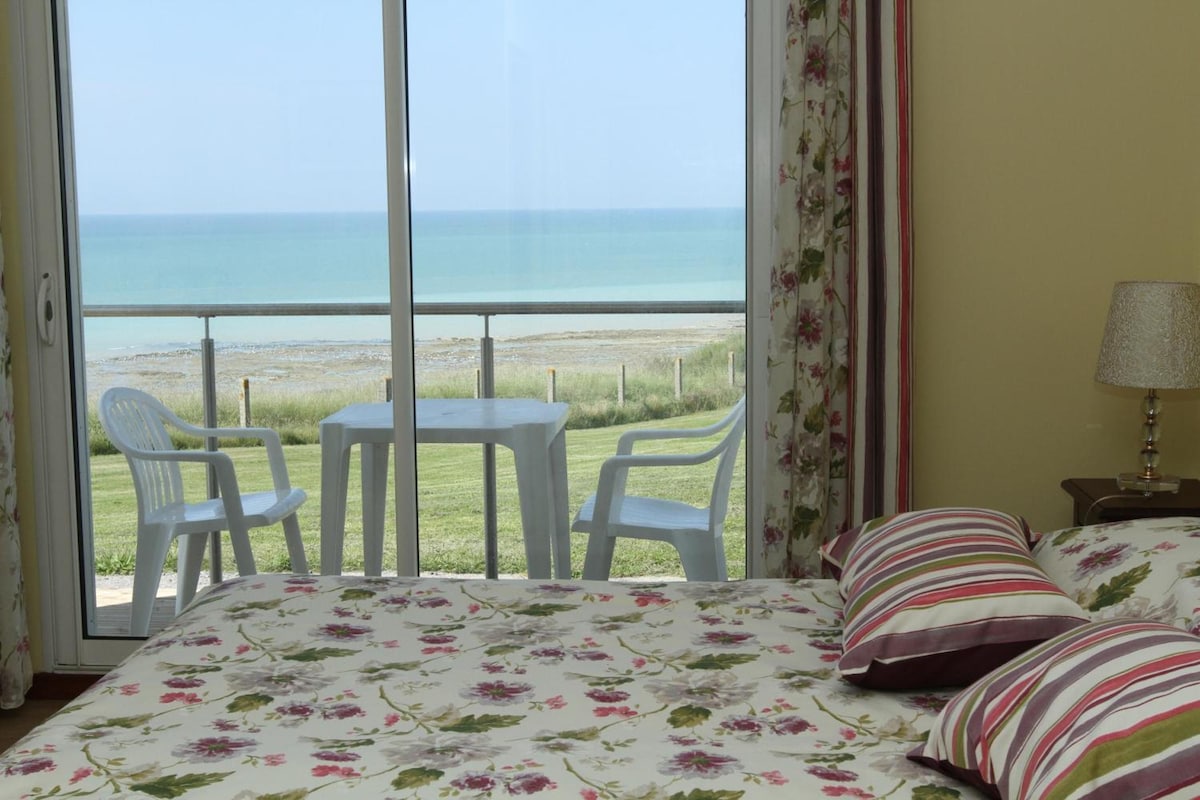 Una habitación con cama, una mesita, dos sillas, una lámpara y vista al mar.