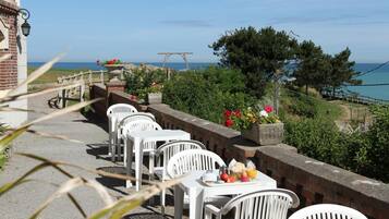 Una terraza frente al mar con sillas y mesas de plástico blanco, un muro de ladrillo y vistas al océano.
