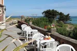 Una terraza frente al mar con sillas y mesas de plástico blanco, un muro de ladrillo y vistas al océano.