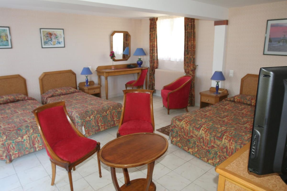 Une chambre d’hôtel avec deux lits, deux chaises rouges, une table en bois, un bureau avec une lampe, une télévision et une fenêtre avec des rideaux.