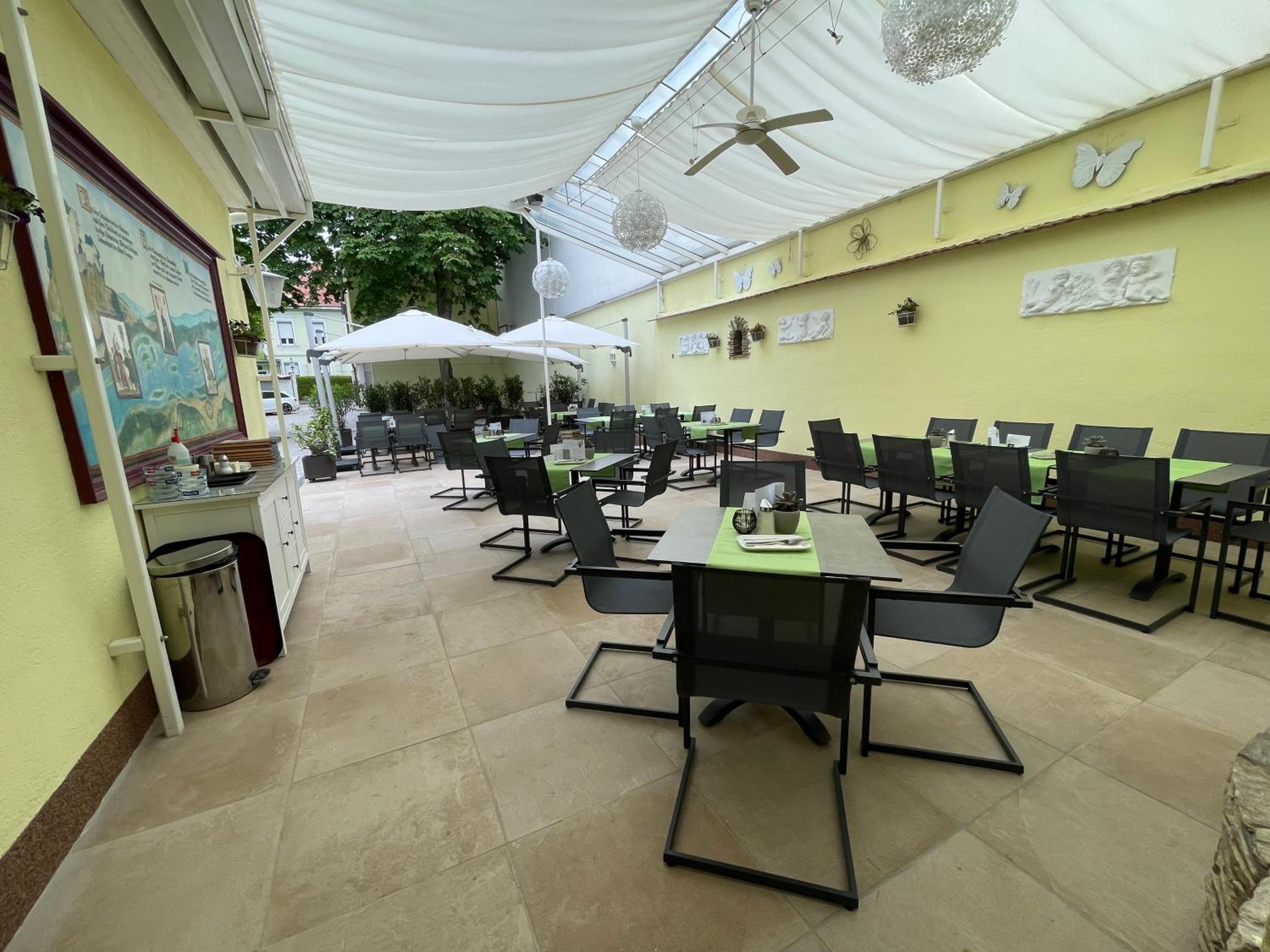Ein überdachter Außenbereich zum Essen mit Tischen und Stühlen, einem Deckenventilator und dekorativen Elementen.